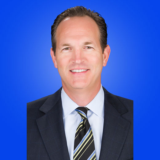 Photo of Brooks Sherman wearing dark gray suit jacket, striped tie, and white shirt on blue background.