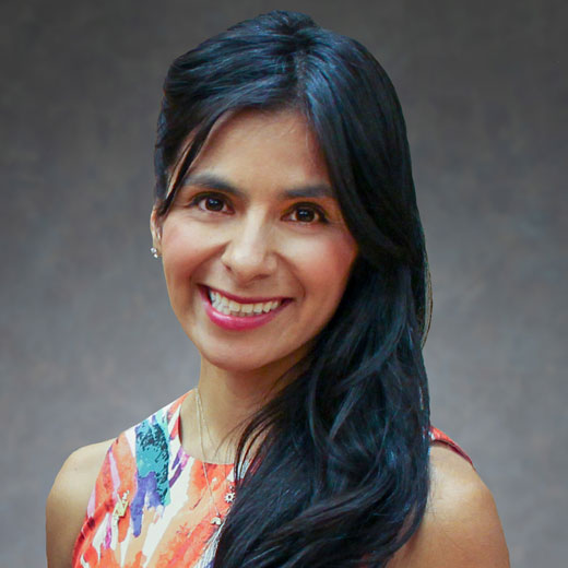 Photo of Mayra Aguirre wearing colorful dress against mottled gray backdrop.