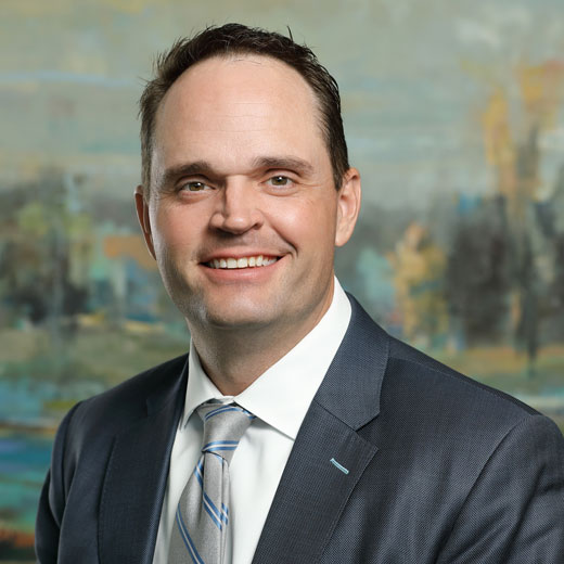 Photo of Sam Huenergardt wearing a gray suit jacket and silver tie with blue stripes, standing in front of a painting.