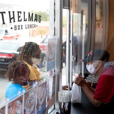 Thelma's Kitchen To Go window with staff inside and two customers wearing face masks outside.