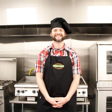 Photo of MCPL Culinary Center Director, Xander Winkel, wearing black chef's hat and apron, standing in front of stainless steel commercial kitchen appliances.