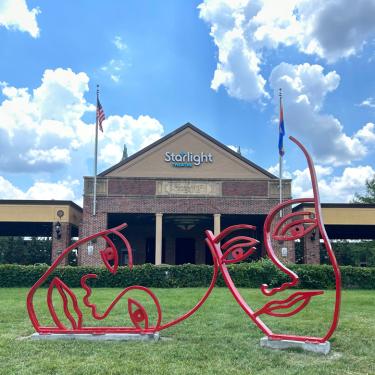Photo of sculpture comprised of two large, abstracted faces, inspired by the traditional theatre masks, made of bent steel tubing painted red, on the lawn in front of Starlight Theatre's box office entrance.